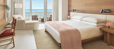 Habitación de hotel con una cama grande, un escritorio con silla, una mesita con sillas y vistas al océano a través de amplias ventanas.
