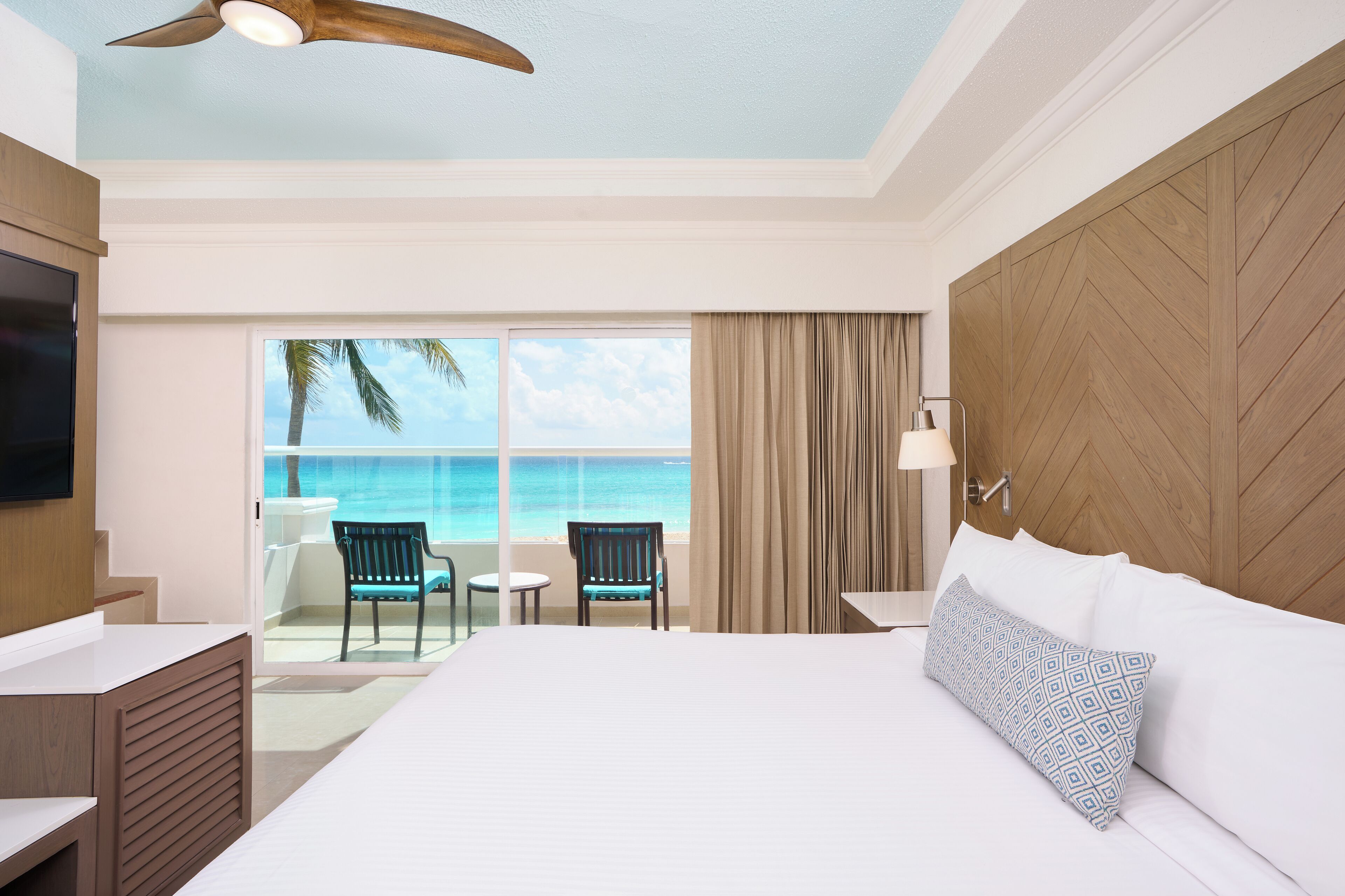 Habitación de hotel con una cama grande, televisión, balcón con vista al océano y cabecero de madera.