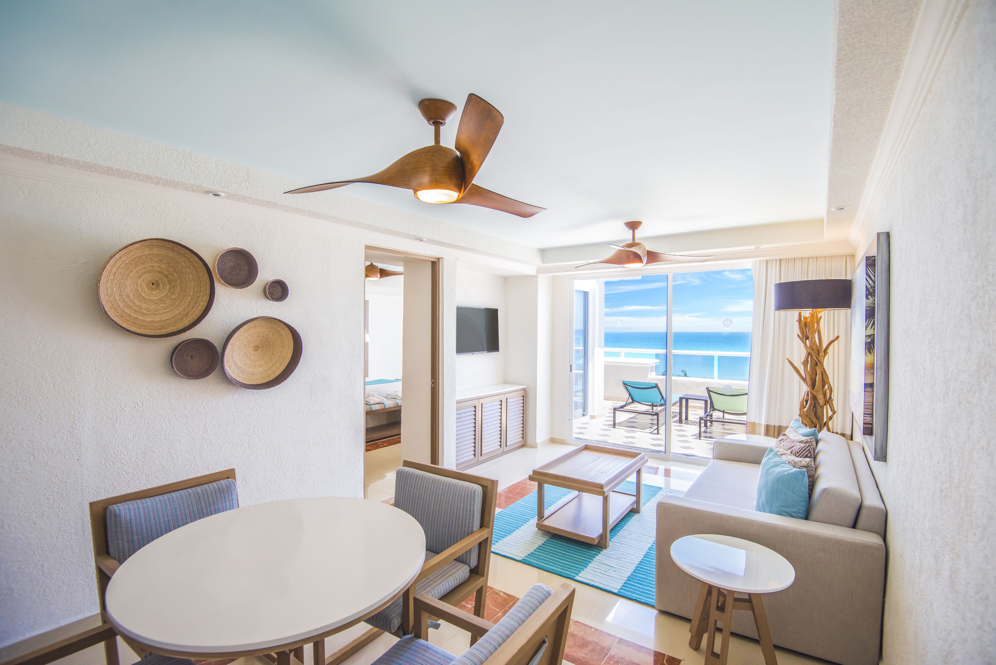 Een ruime woonkamer met een ronde tafel, banken en een plafondventilator. Er zijn decoratieve wandplaten en een uitzicht op de oceaan door grote ramen.