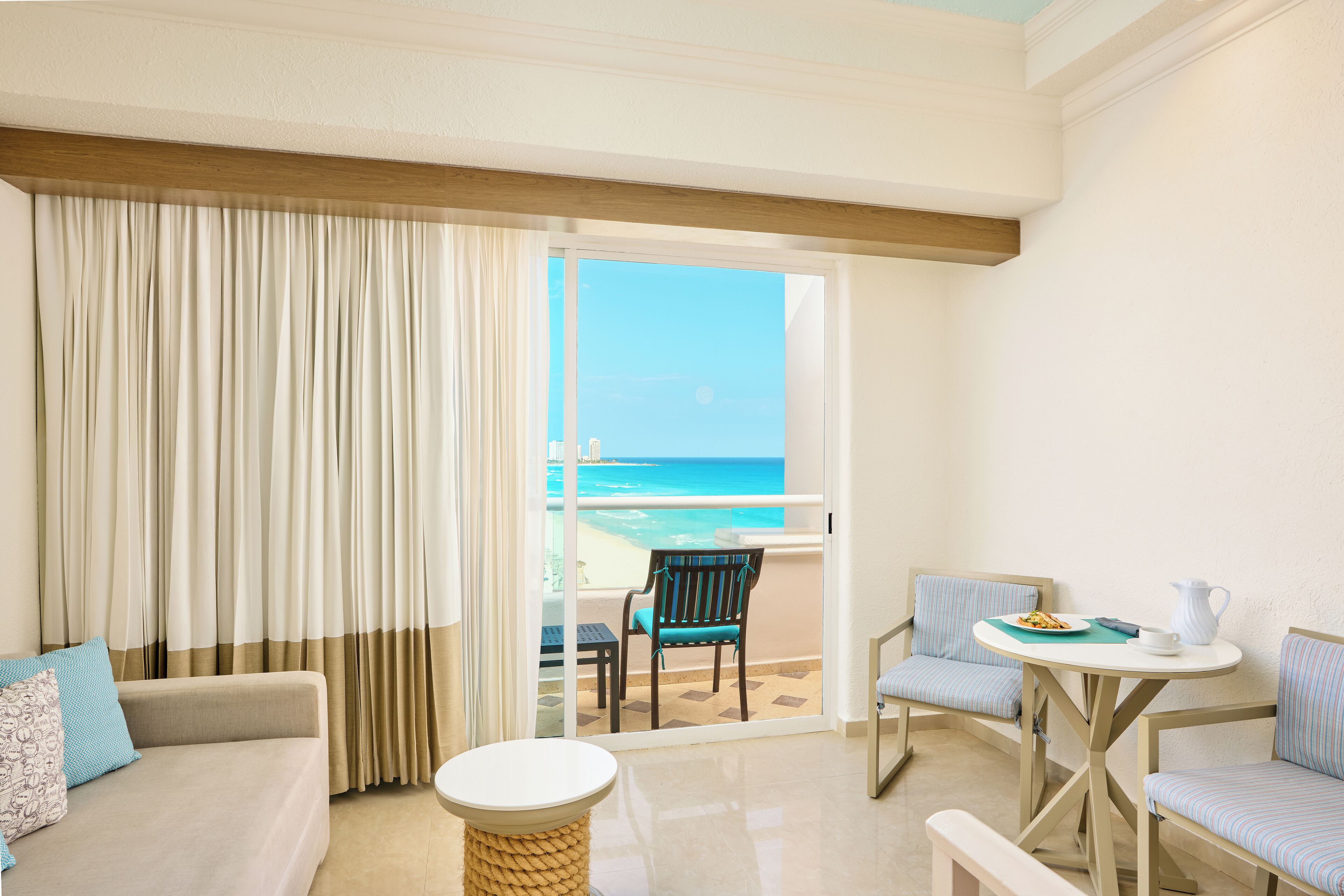 Uma varanda com vista para a praia, um sofá, duas cadeiras listradas, uma mesa pequena e um banquinho de vime.