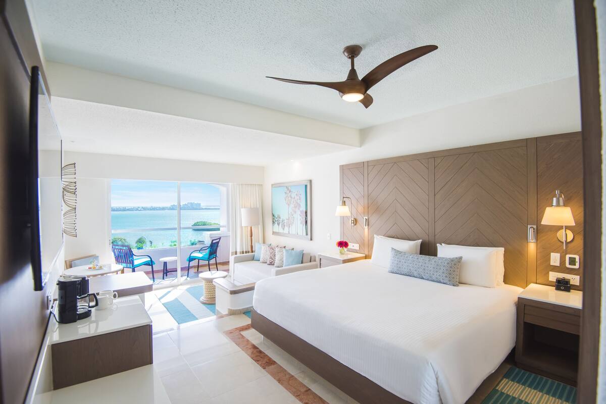 A hotel room with a large bed, a ceiling fan, a view of the sea, a dining area with a table and chairs, and a kitchenette.