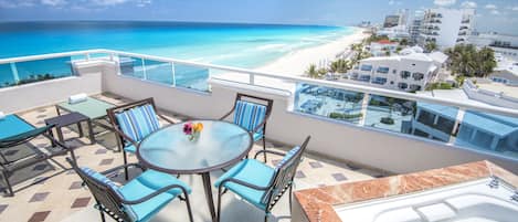 Un balcón con mesa de vidrio, sillas y bañera de hidromasaje con vistas a una playa y edificios de un complejo turístico.