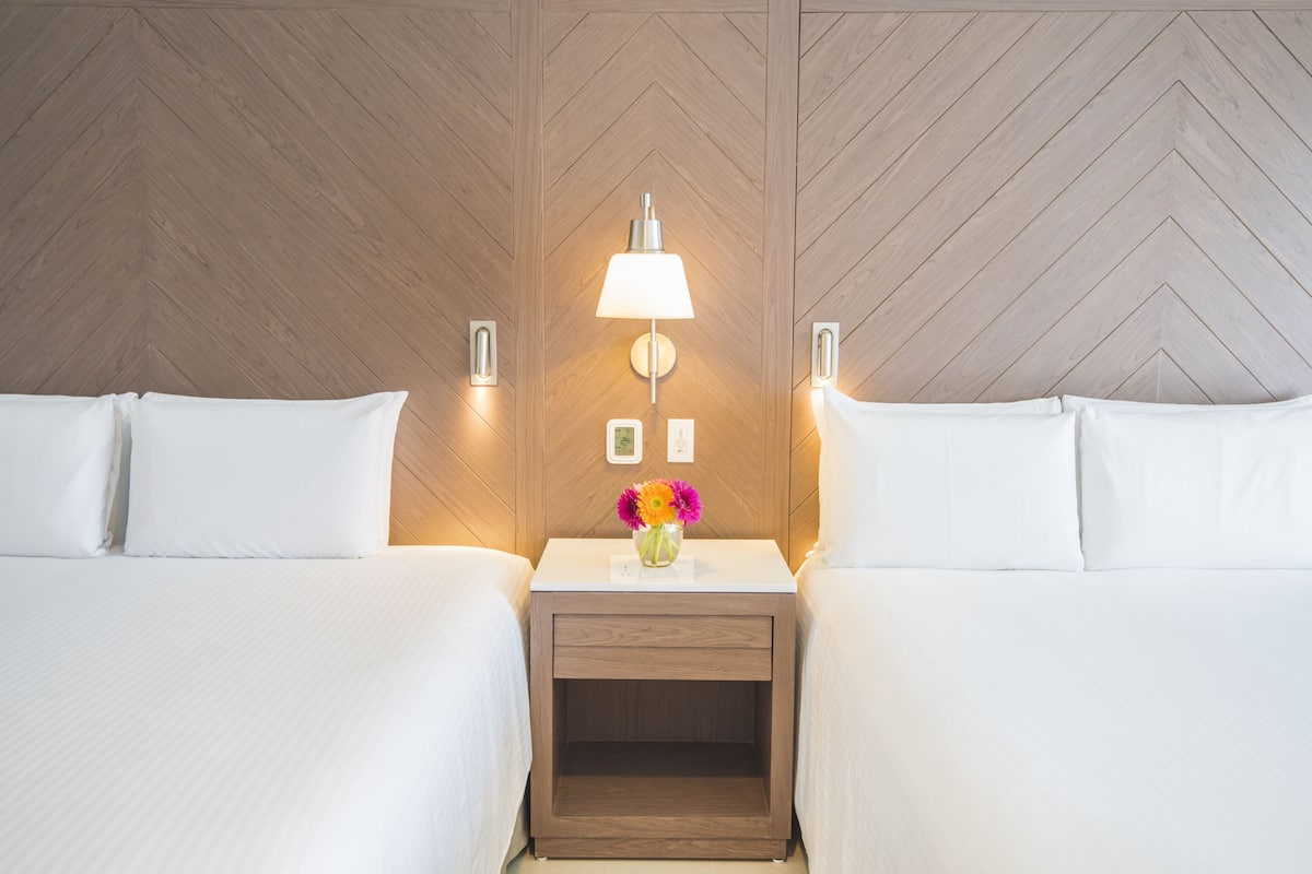 A hotel room with two beds, a nightstand with a lamp, and a flower vase.