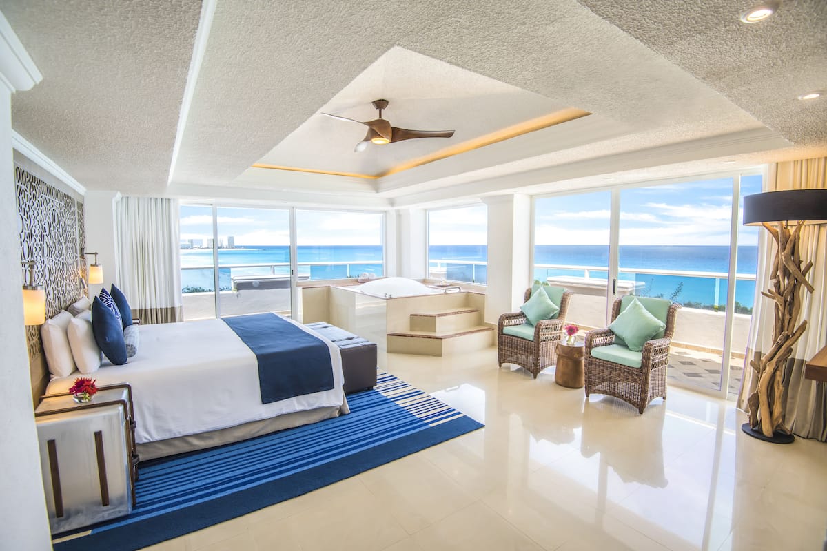 A spacious hotel room with a large bed, a balcony with a view of the ocean, and a seating area with wicker furniture.