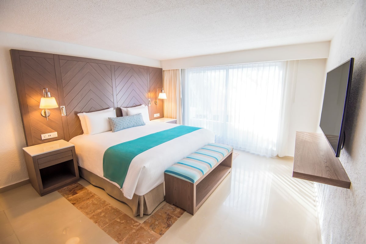 A modern hotel room with a large bed, bedside lamps, a flat-screen TV, and a large window with curtains.