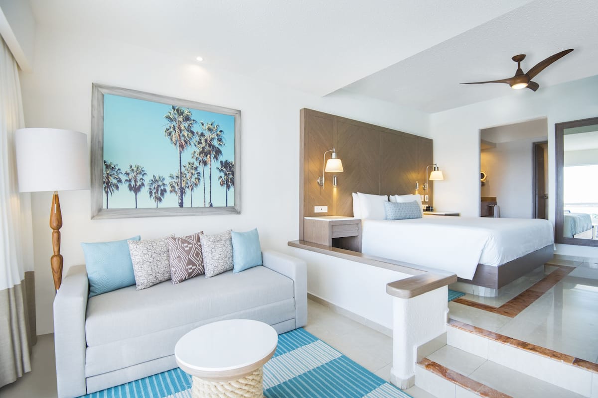 A modern hotel room with a bed, a sofa, a small table, and a painting of palm trees.