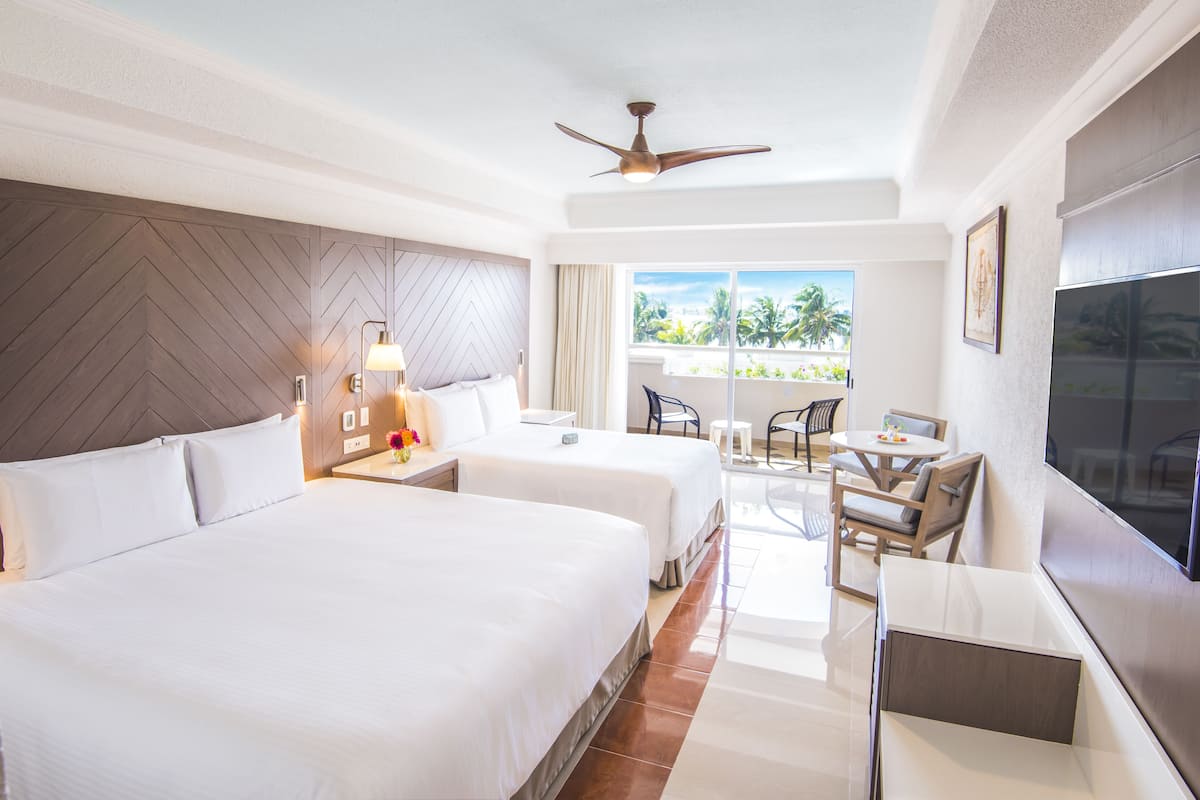 A hotel room with two beds, a ceiling fan, a TV, a balcony with a view of palm trees, and a dining area with chairs and a table.