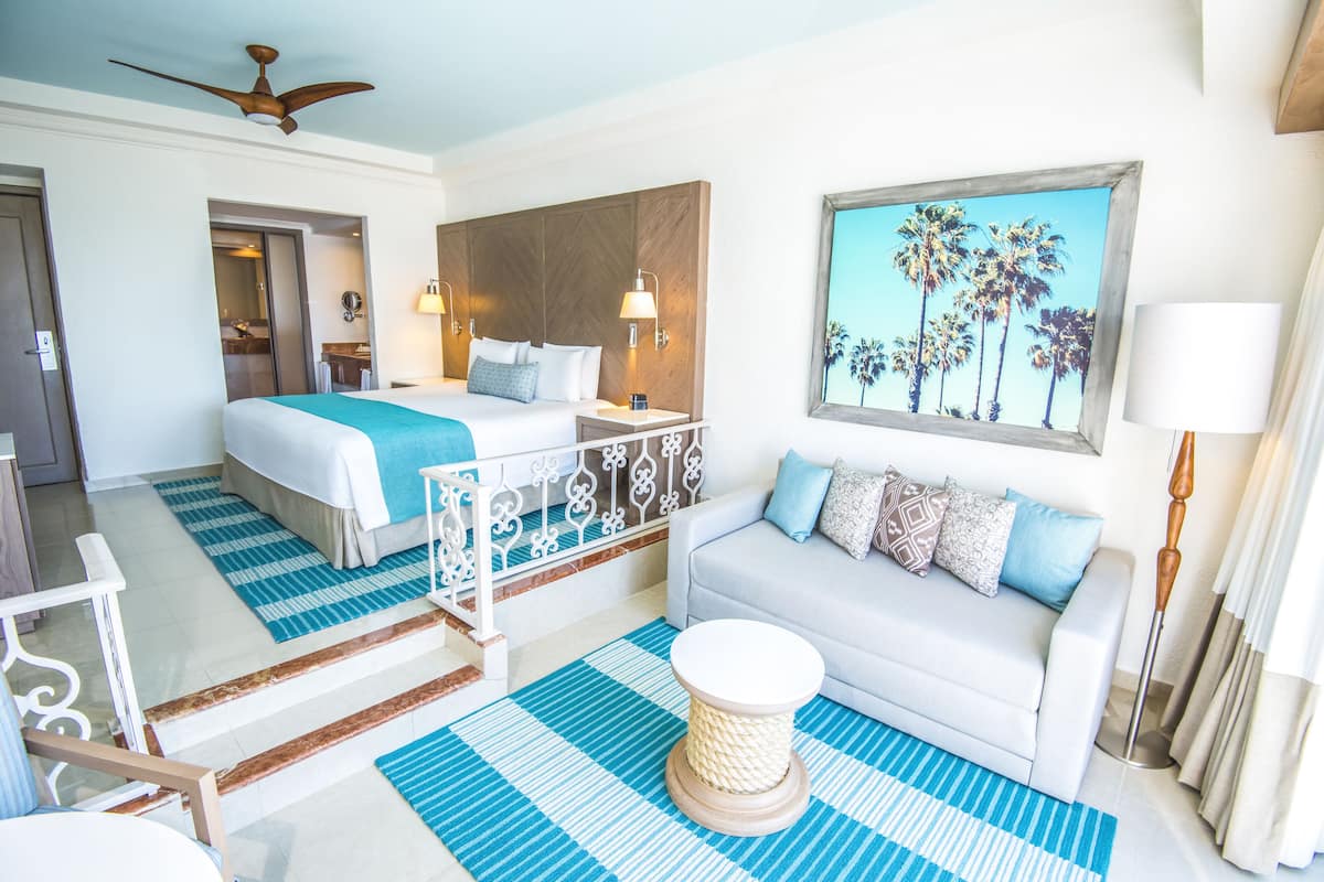 A hotel room with a bed, a sofa, a painting of palm trees, a ceiling fan, and a window with curtains.