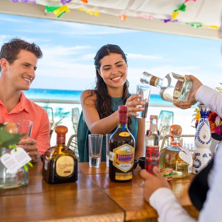 A group of people at a table with various bottles and glasses, likely a bar setting, with a view of the ocean in the background.