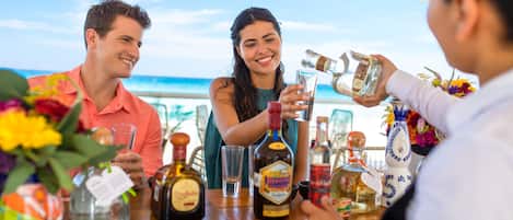 A group of people at a table with various bottles and glasses, likely a bar setting, with a view of the ocean in the background.