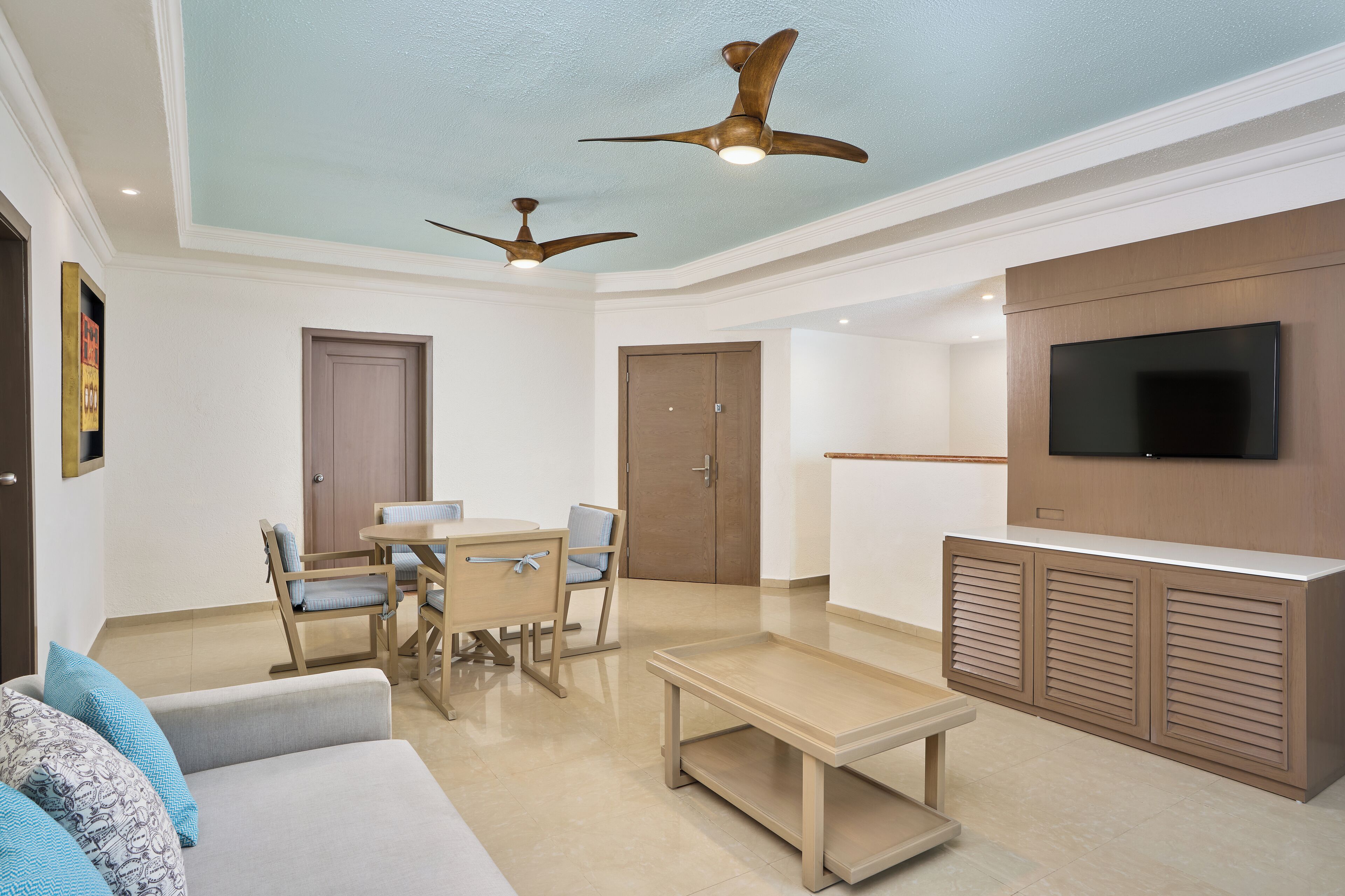 A modern living room with a sofa, dining table, chairs, ceiling fans, and a TV mounted on a wooden cabinet.