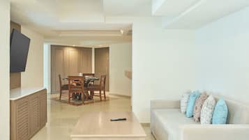A modern living room with a flat-screen TV mounted on the wall, a wooden dining table with chairs, a light-colored sofa with patterned cushions, and a coffee table with a remote control.