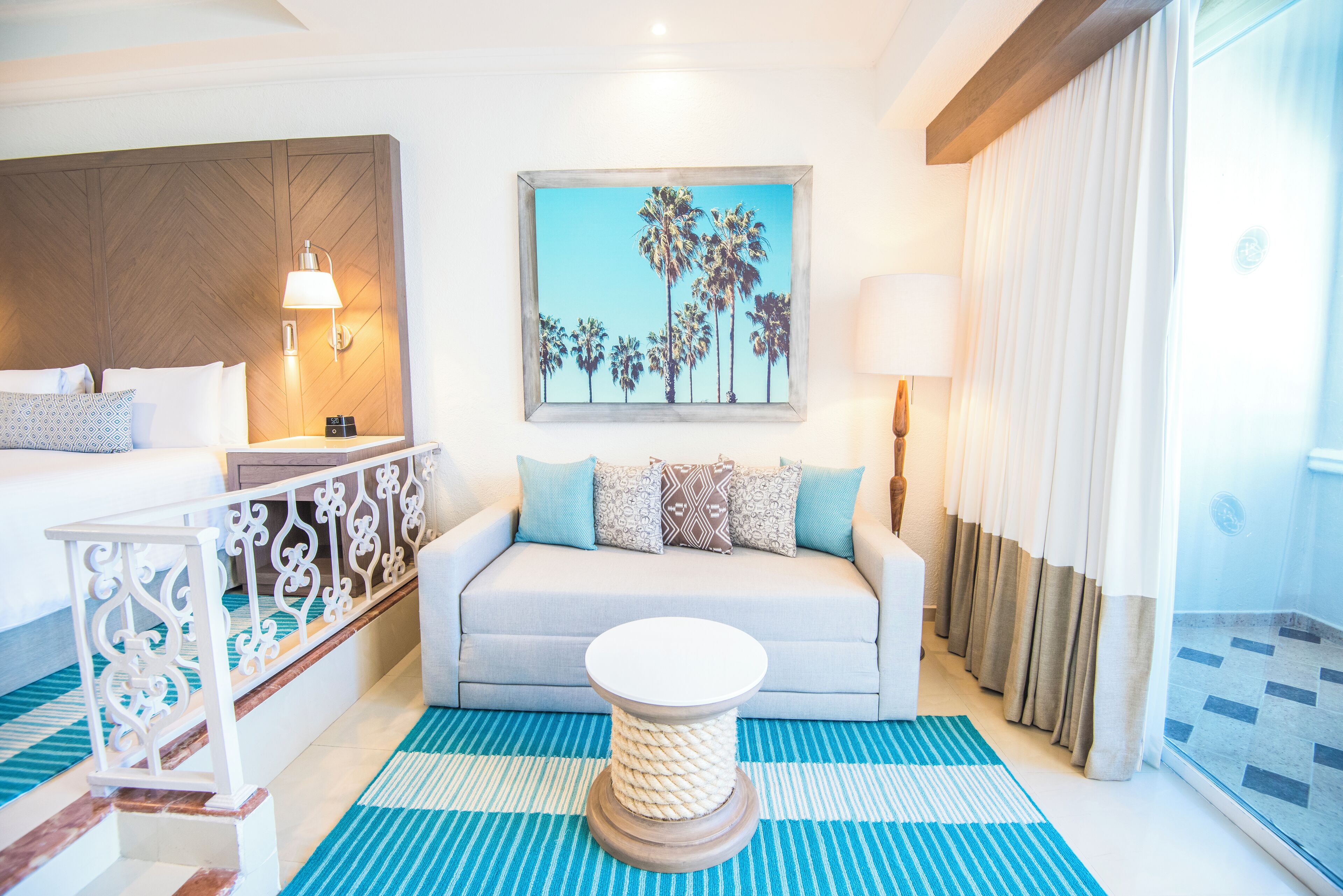 Habitación de hotel con cama, sofá, mesa de centro, pintura de palmeras y ventana con cortinas.