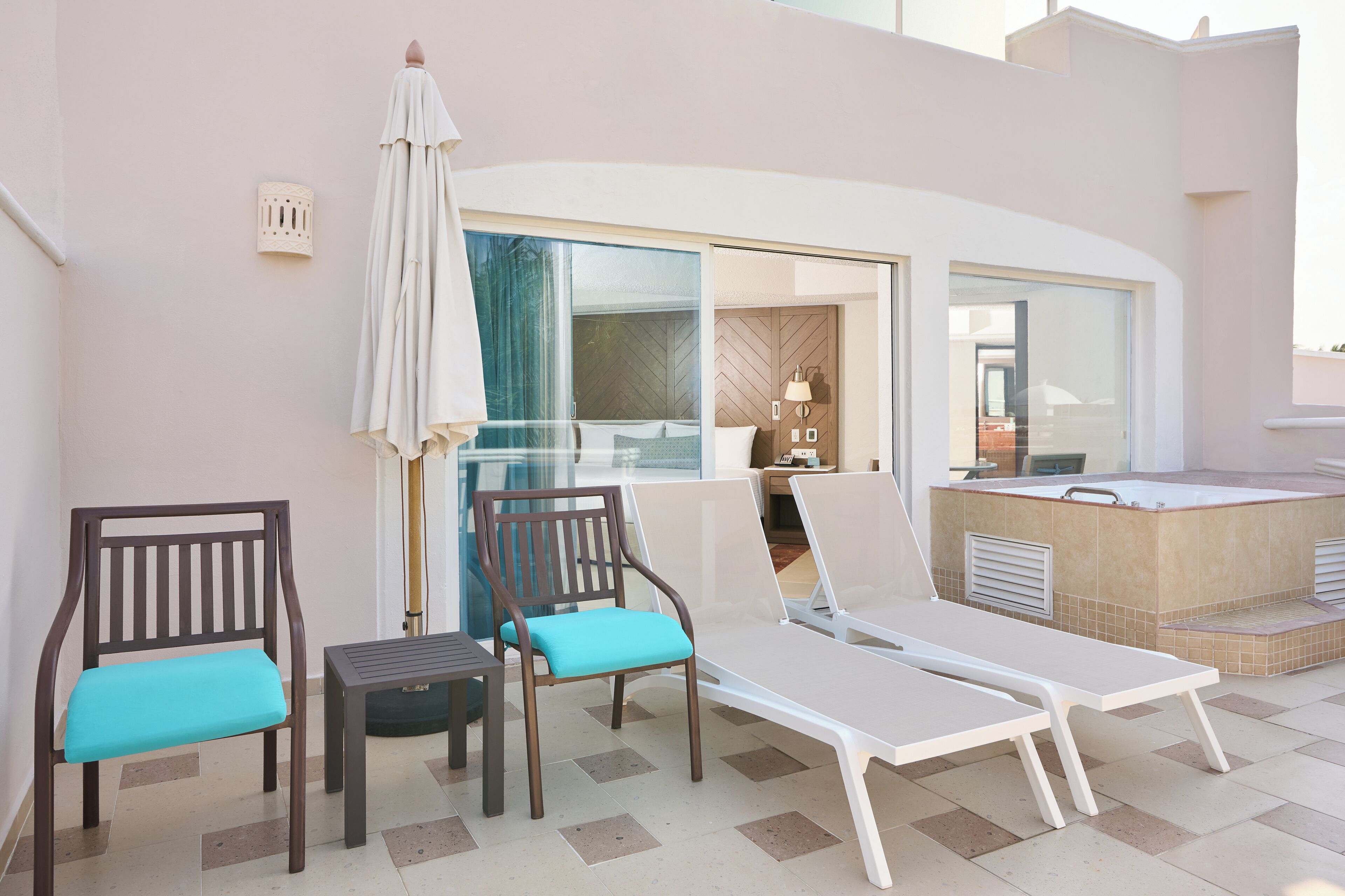 A sunlit outdoor seating area with lounge chairs and a table, a glass door leading to a room, and a small hot tub.