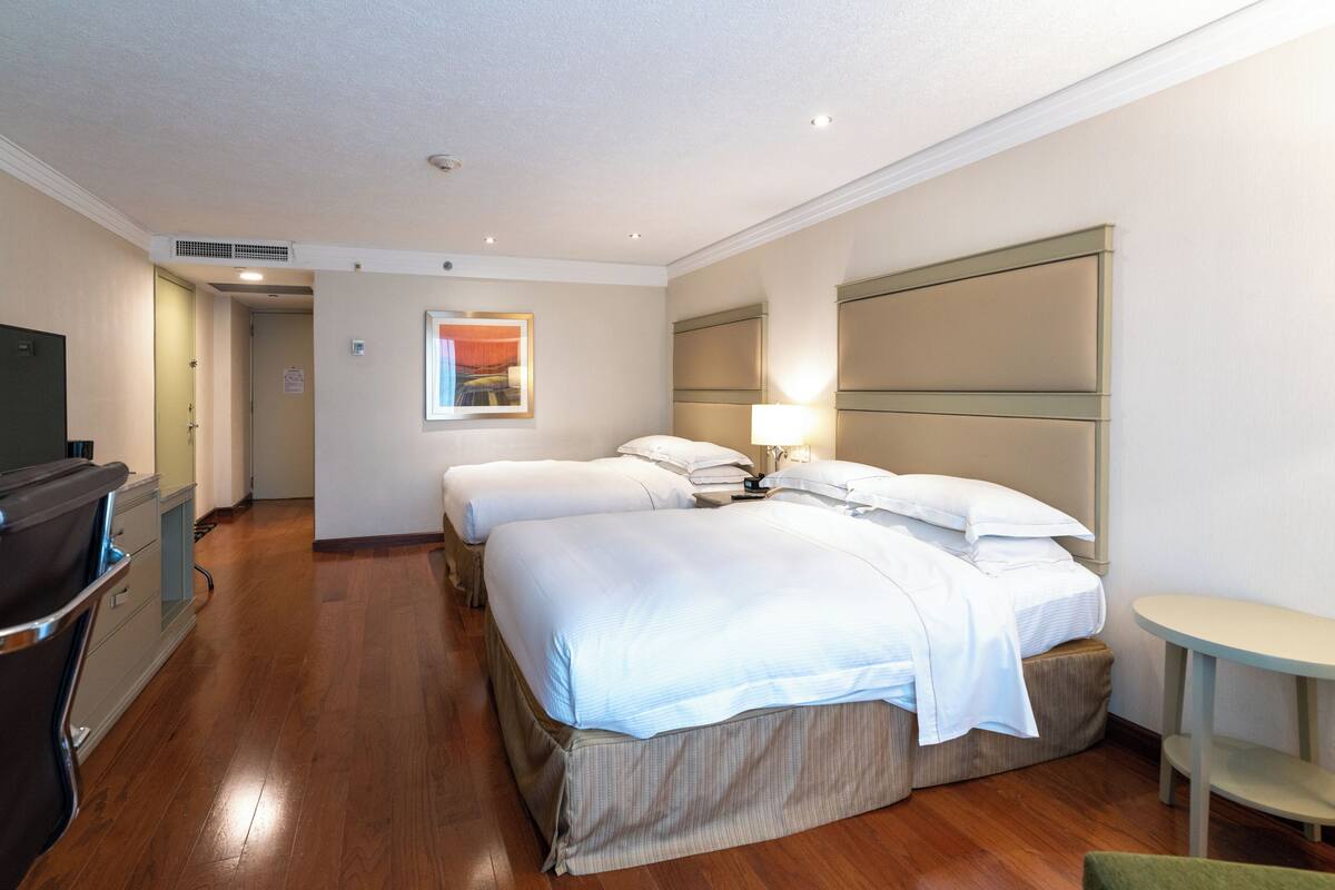 A hotel room with a wooden floor, two beds with white linens, a desk with a chair, and a small table.