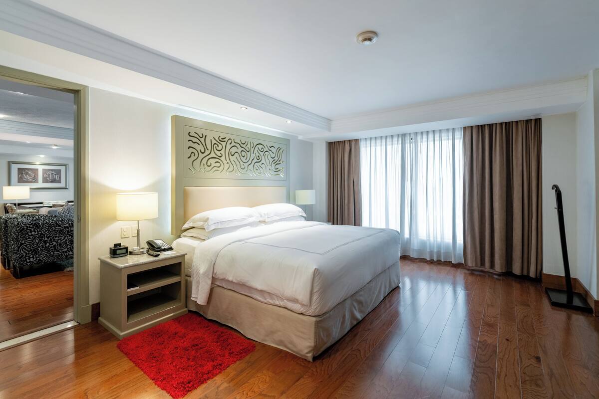 A hotel room with a bed, bedside table, lamp, and a red rug.