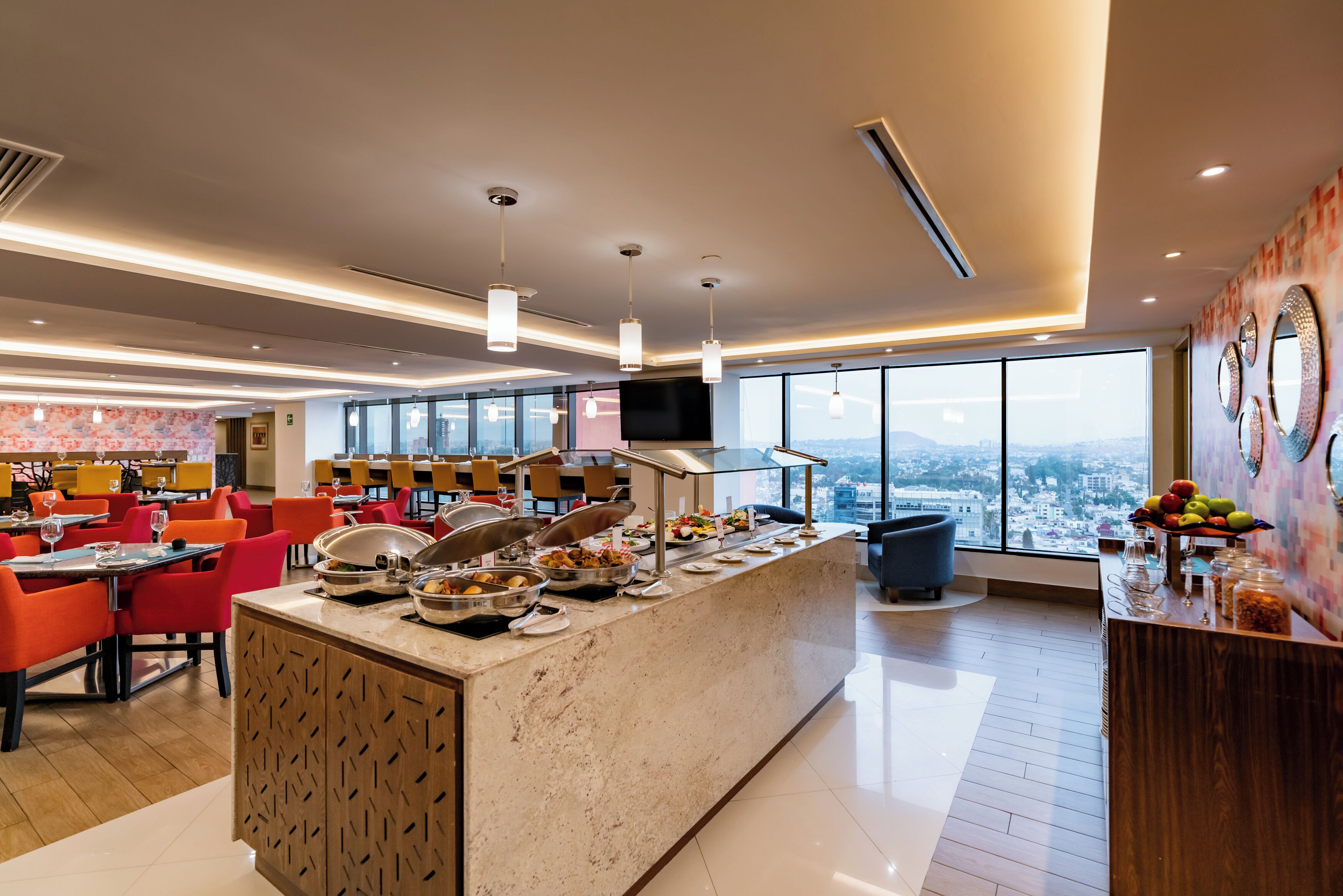Comedor moderno de hotel con bufé, asientos y un amplio ventanal con vistas a la ciudad.