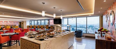 Comedor moderno de hotel con bufé, asientos y un amplio ventanal con vistas a la ciudad.
