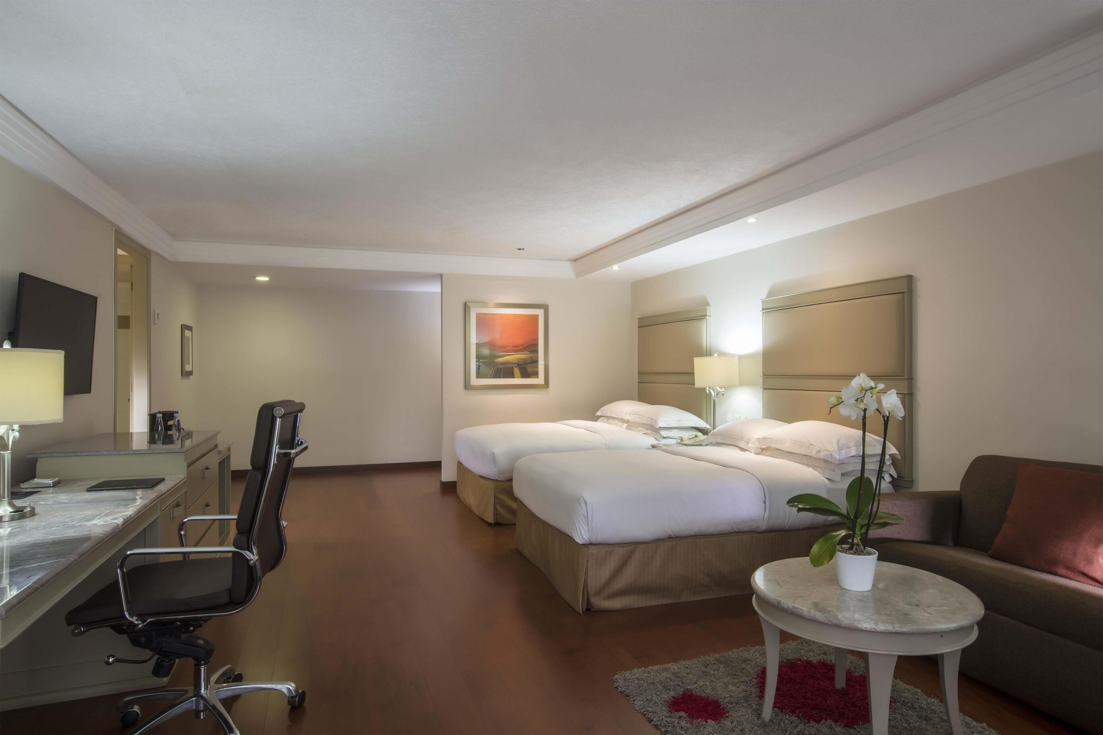 A hotel room with a desk, two beds, a chair, a small table with a plant, and a couch.