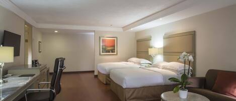 A hotel room with a desk, two beds, a chair, a small table with a plant, and a couch.