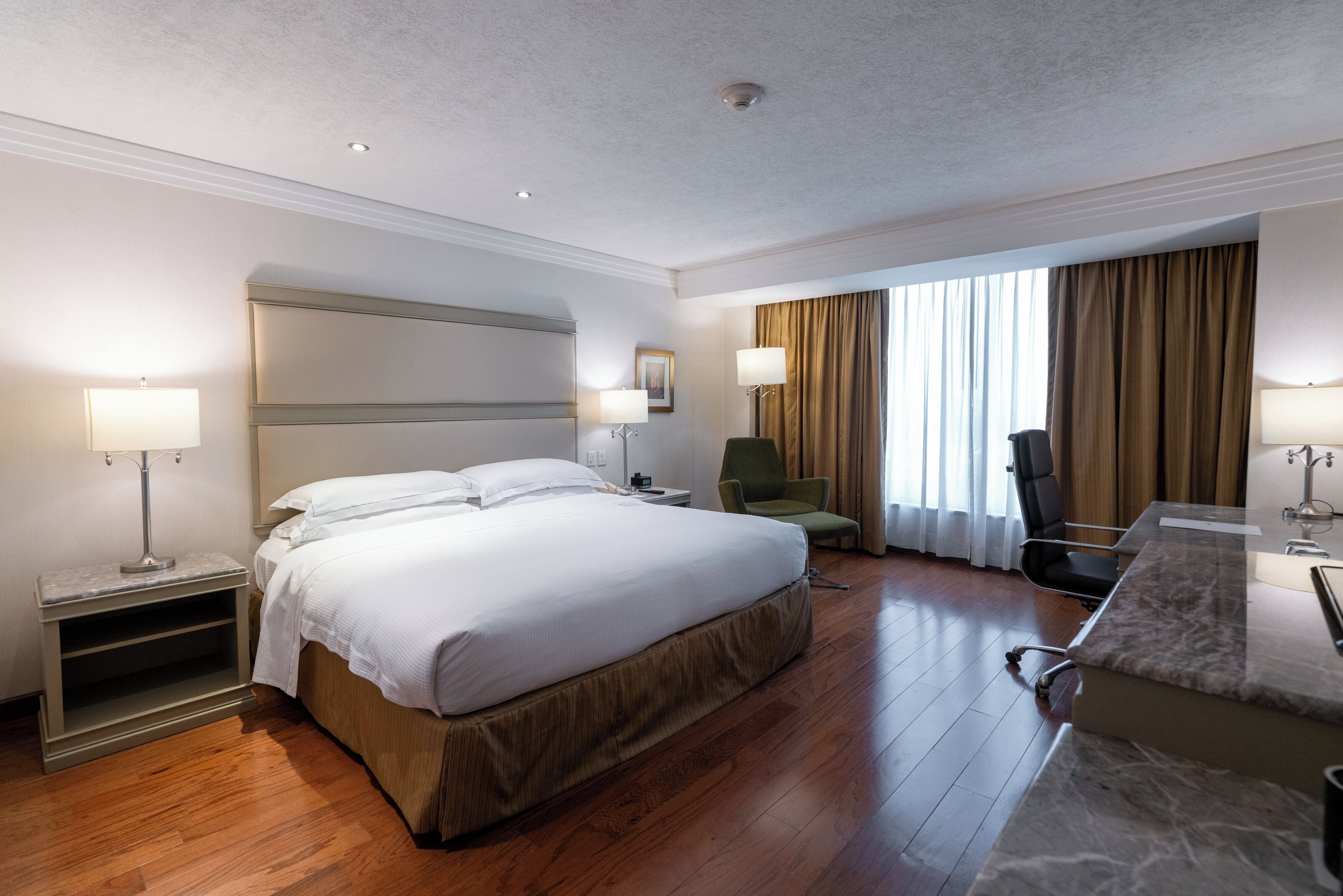 A hotel room with a large bed, a desk, a chair, a lamp, and a window with curtains.