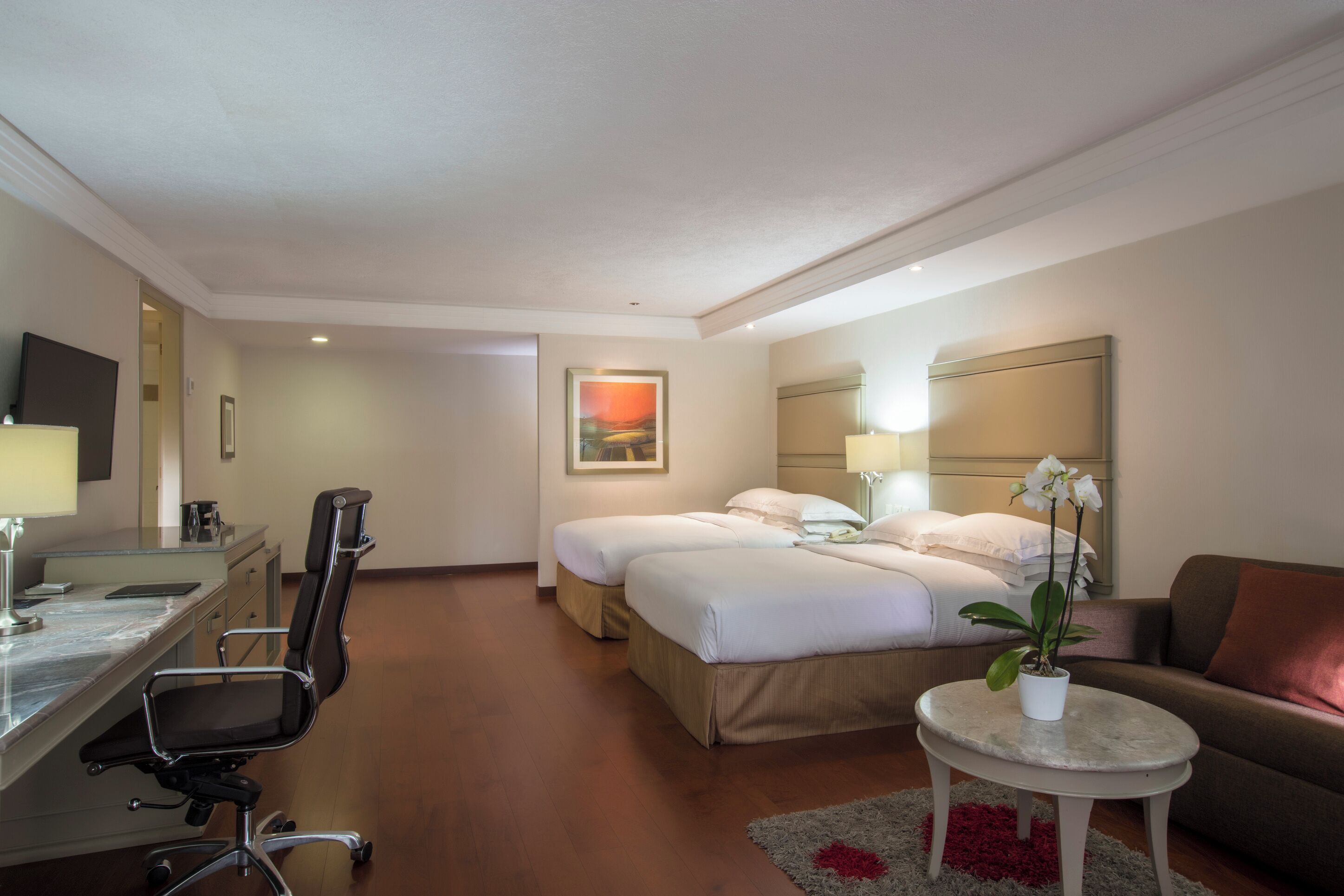 Habitación de hotel con escritorio, dos camas, una silla, una mesita con una planta y un sofá.