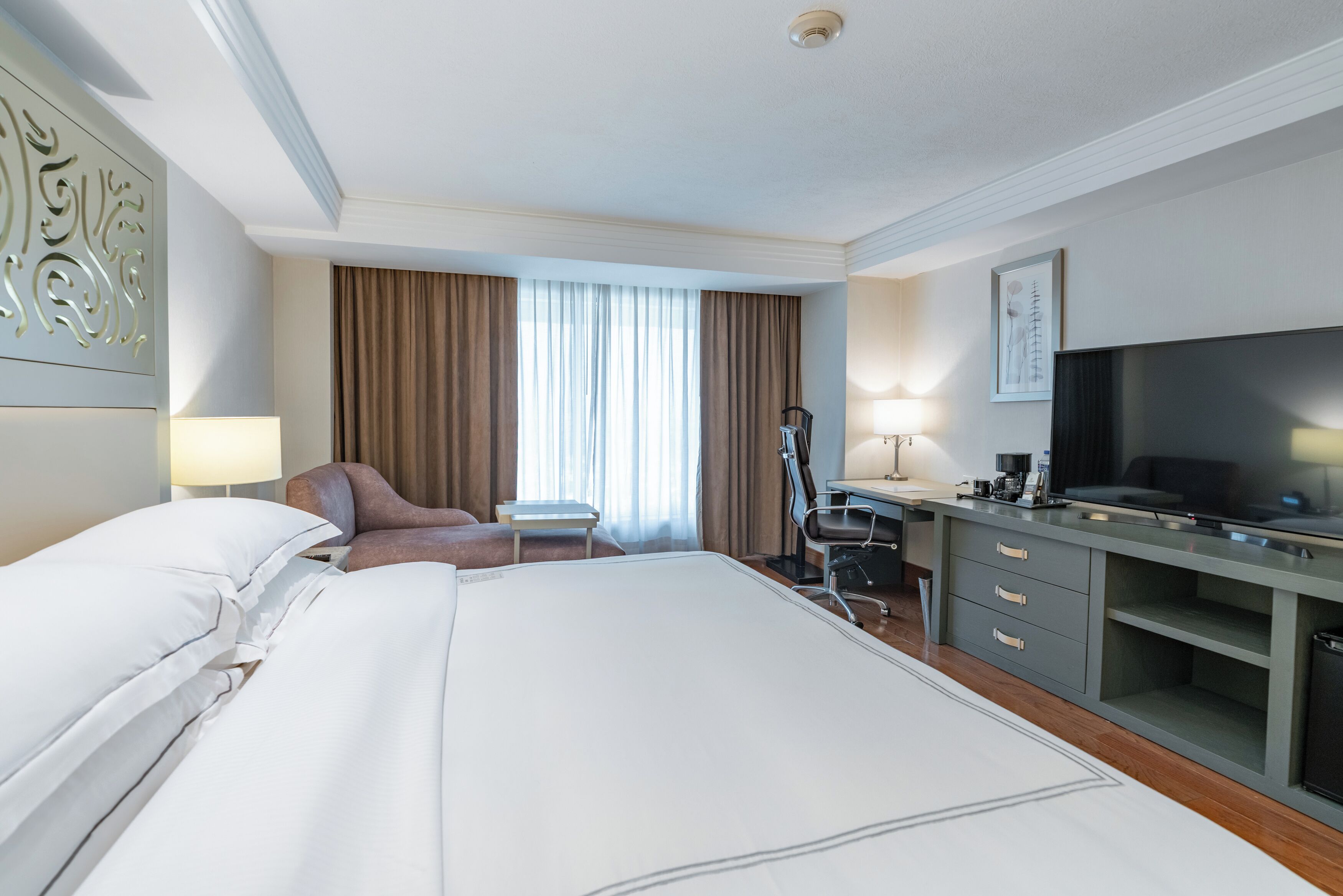 Una habitación de hotel con una cama grande, un escritorio, una silla, un televisor y una ventana con cortinas.