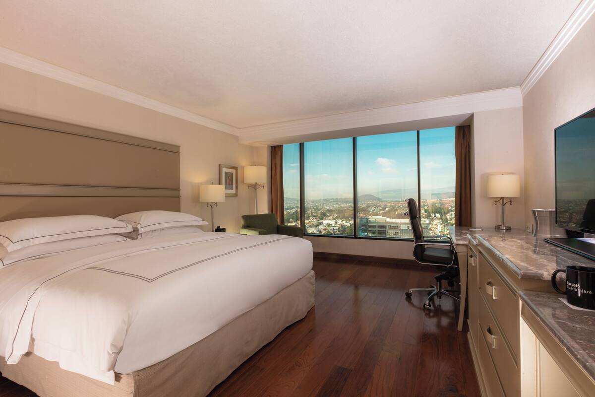 A hotel room with a large bed, a desk, a chair, a TV, and a view of the city.