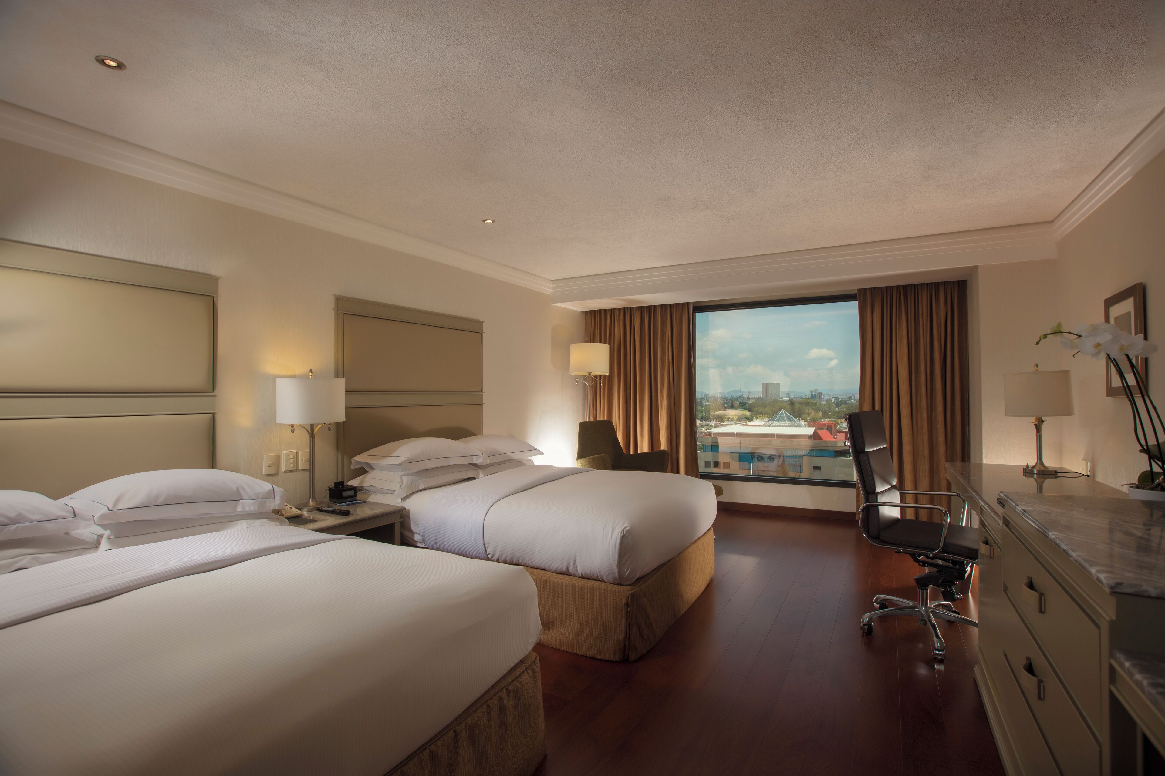 Quarto de hotel com duas camas, uma mesa, uma cadeira e vista para a cidade.