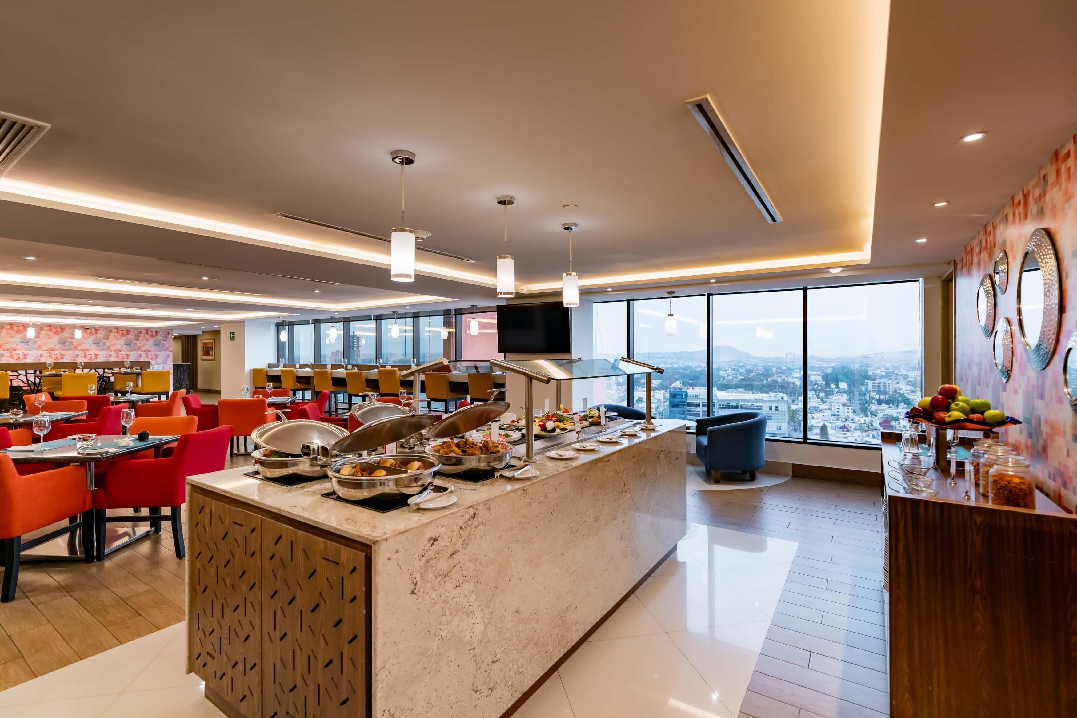 Espaço de refeições moderno em hotel, com buffet, pé-direito alto e grandes janelas com vista para a cidade.