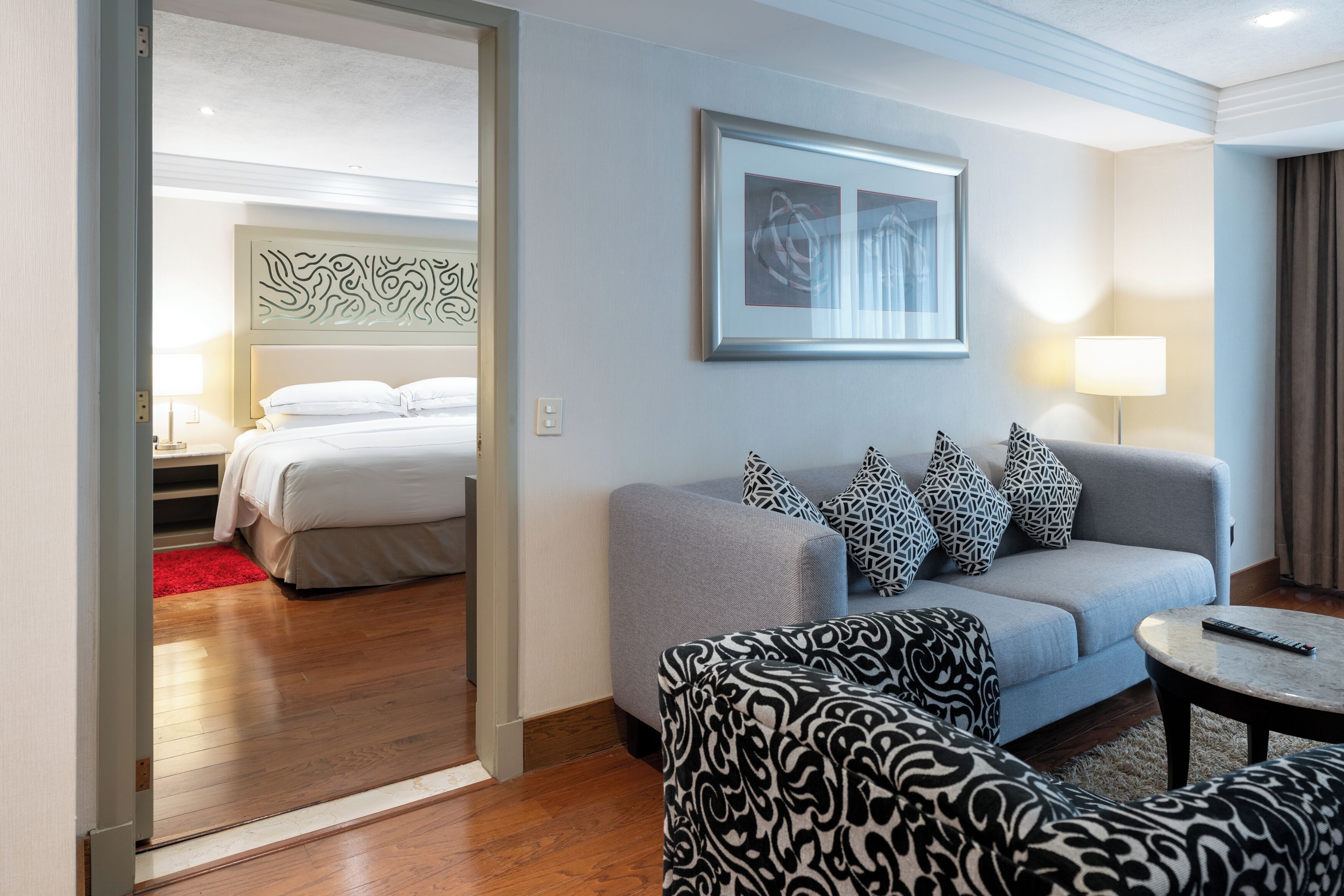 A hotel room with a bed, a grey sofa, patterned cushions, a remote control on a side table, and a framed artwork on the wall.
