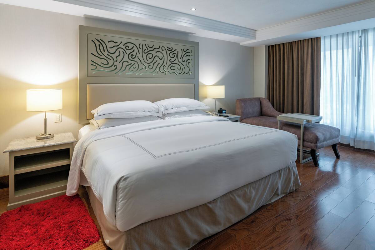 A hotel room with a large bed, two bedside tables, a chair, a desk, a red rug, and a wall art piece.