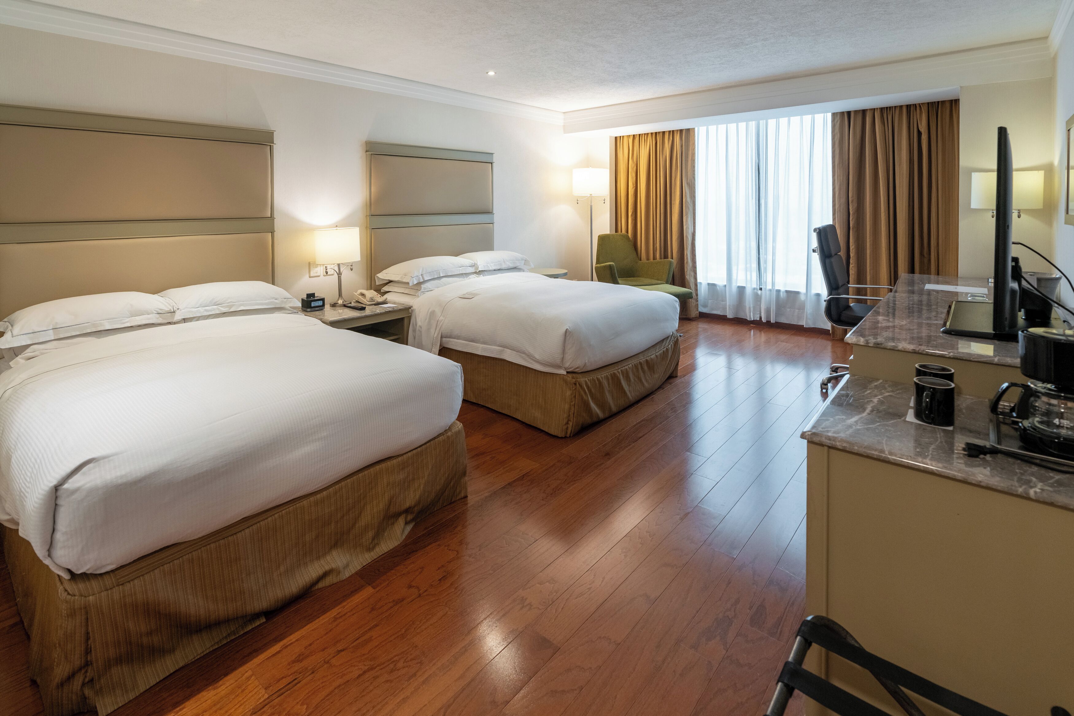 Habitación de hotel con dos camas, un escritorio, una silla, un televisor y un ventanal con cortinas.