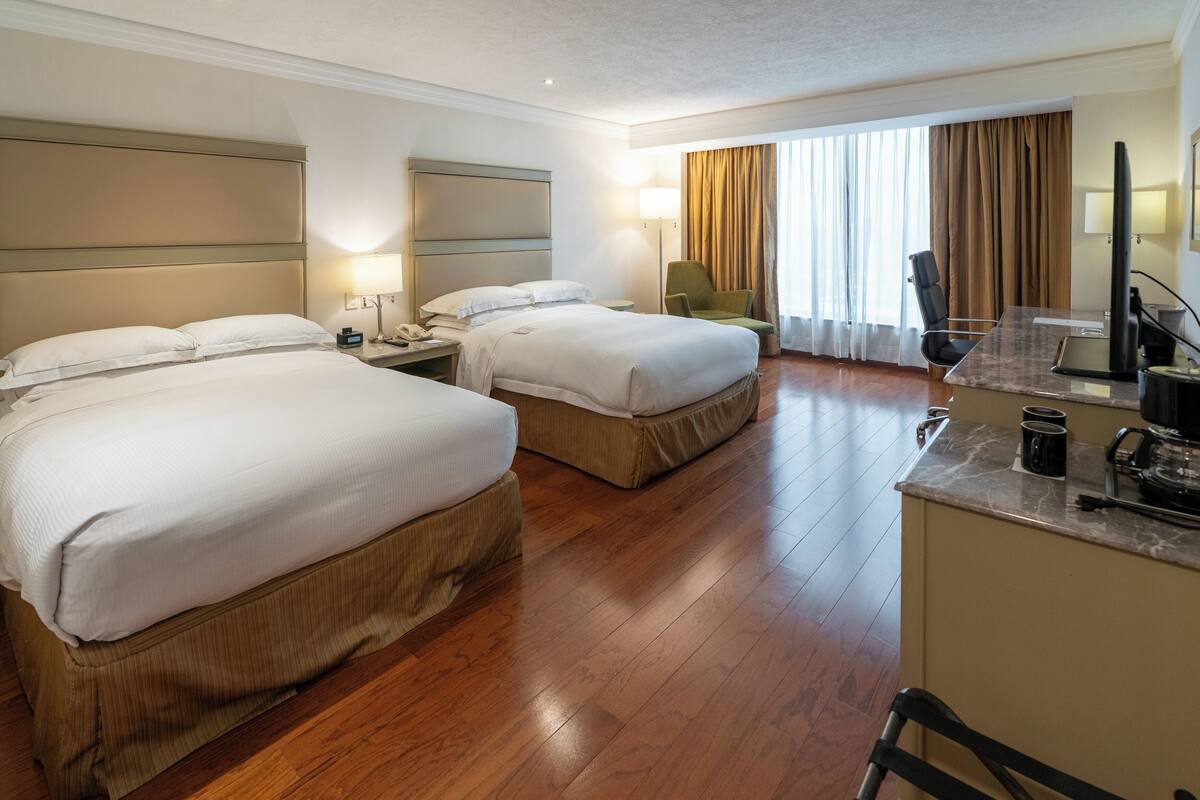 A hotel room with two beds, a desk, a chair, a TV, and a large window with curtains.