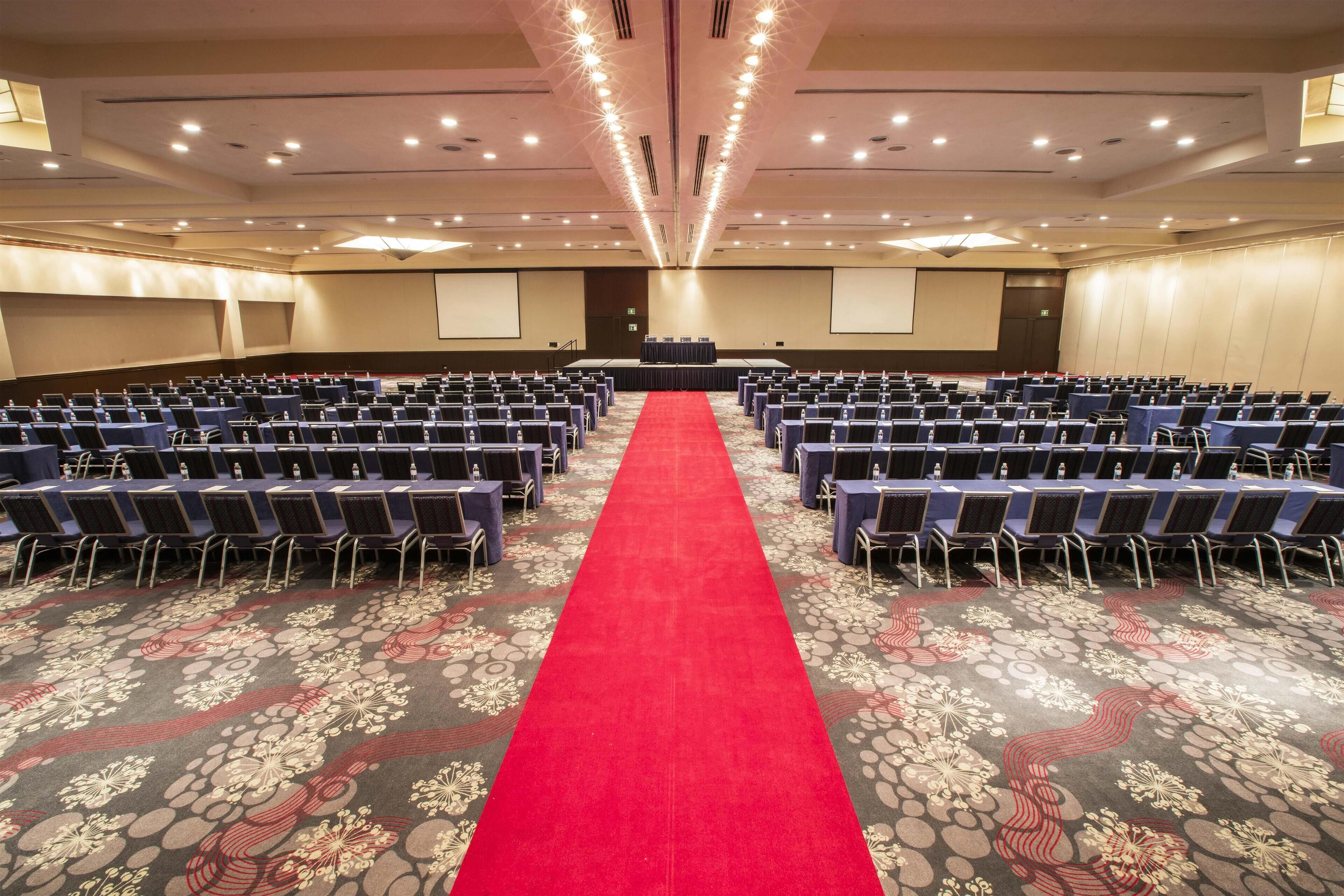A conference room with rows of chairs, a red carpet, and a projector screen.