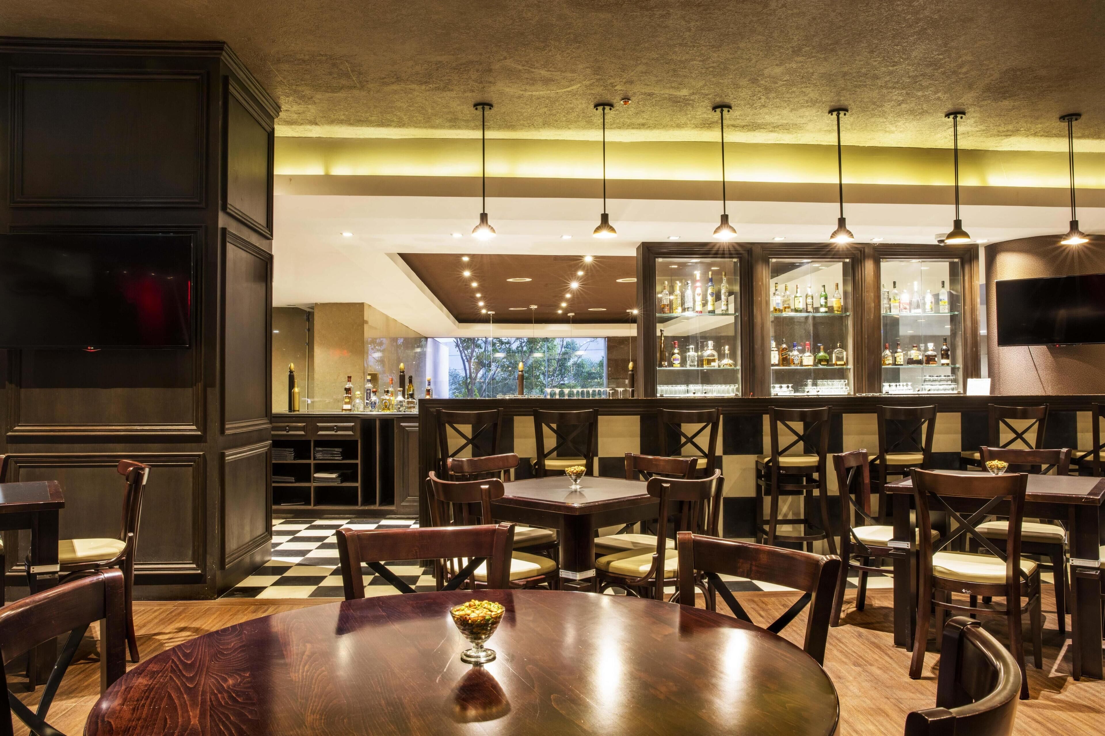 Un restaurante con mesas y sillas de madera, una zona de bar con estantes llenos de botellas y un televisor colgado en la pared.