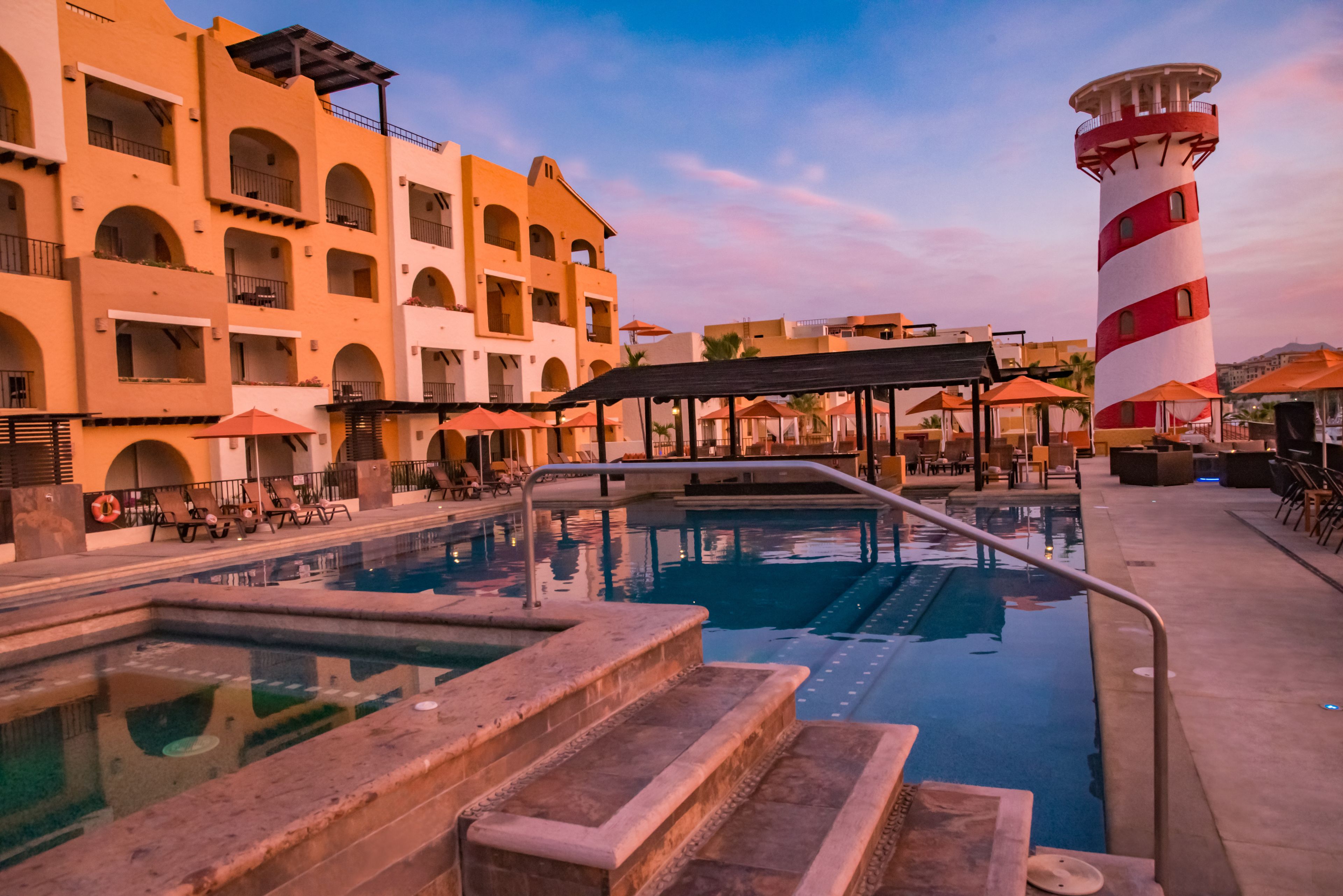 A pool area with steps leading into the water, surrounded by buildings with arched windows and balconies, and a lighthouse in the background.