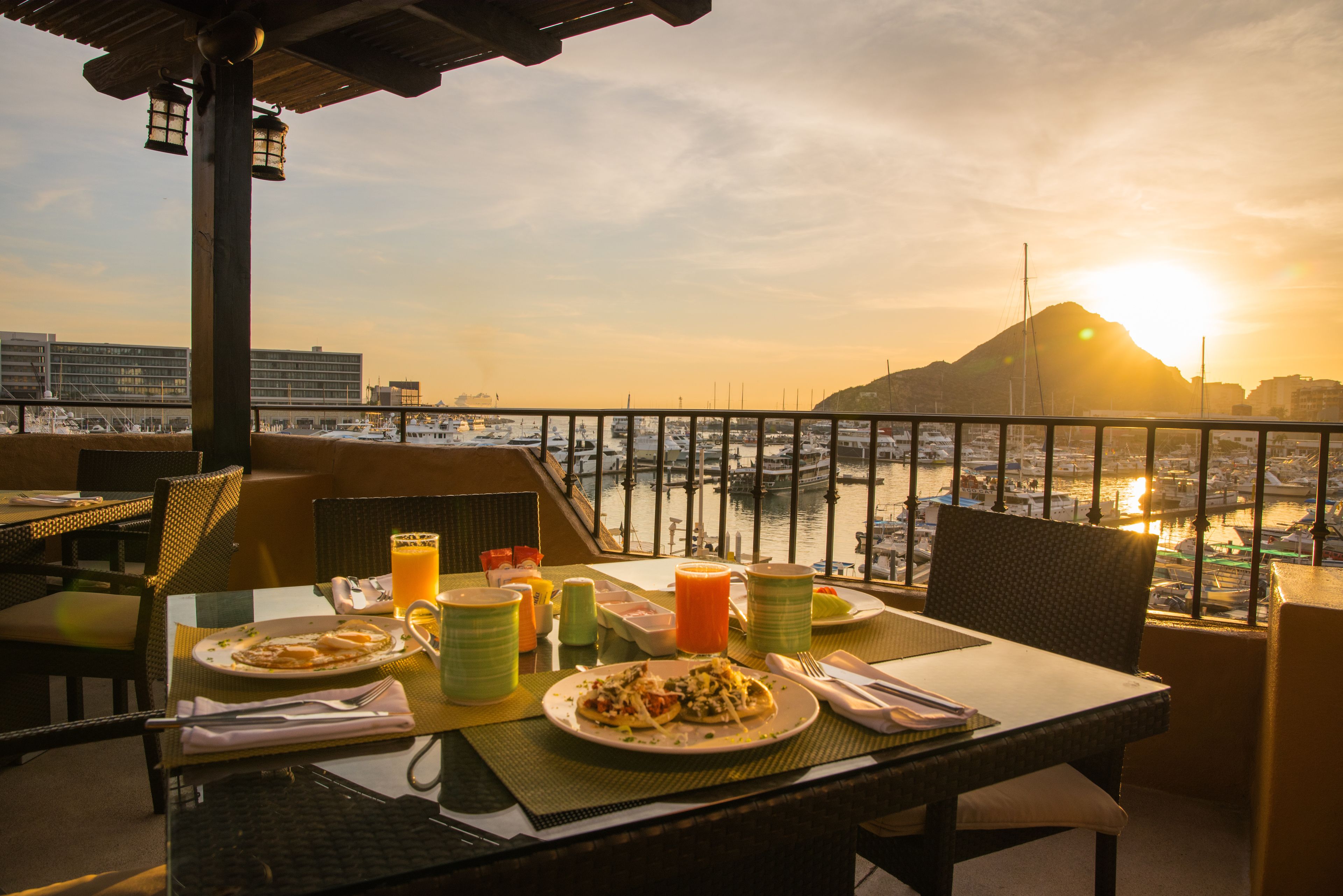 Una mesa de comedor con platos de comida, vasos y cubiertos, con vista a un puerto deportivo con barcos y una montaña al fondo.