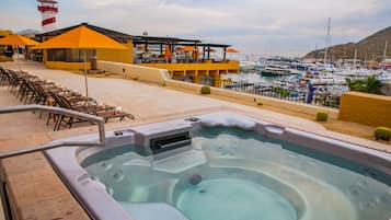 Een bubbelbadgedeelte met loungebedden en parasols, met uitzicht op een jachthaven met boten en een berg op de achtergrond.