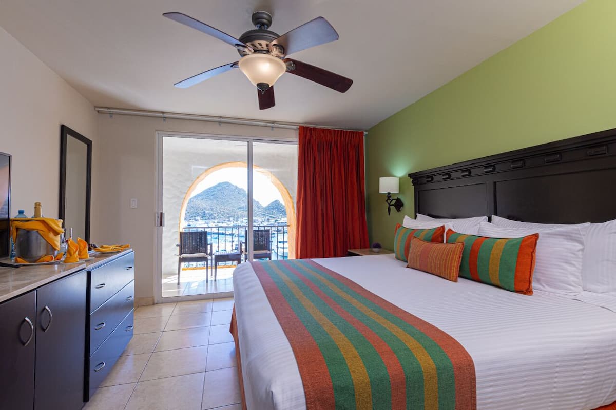 Habitación de hotel con cama, ventilador de techo, mesita de noche y vista al mar a través de una puerta de balcón.