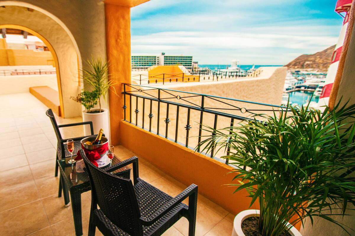 Un balcón con una mesa para dos, una planta en maceta y vista a la playa y edificios.