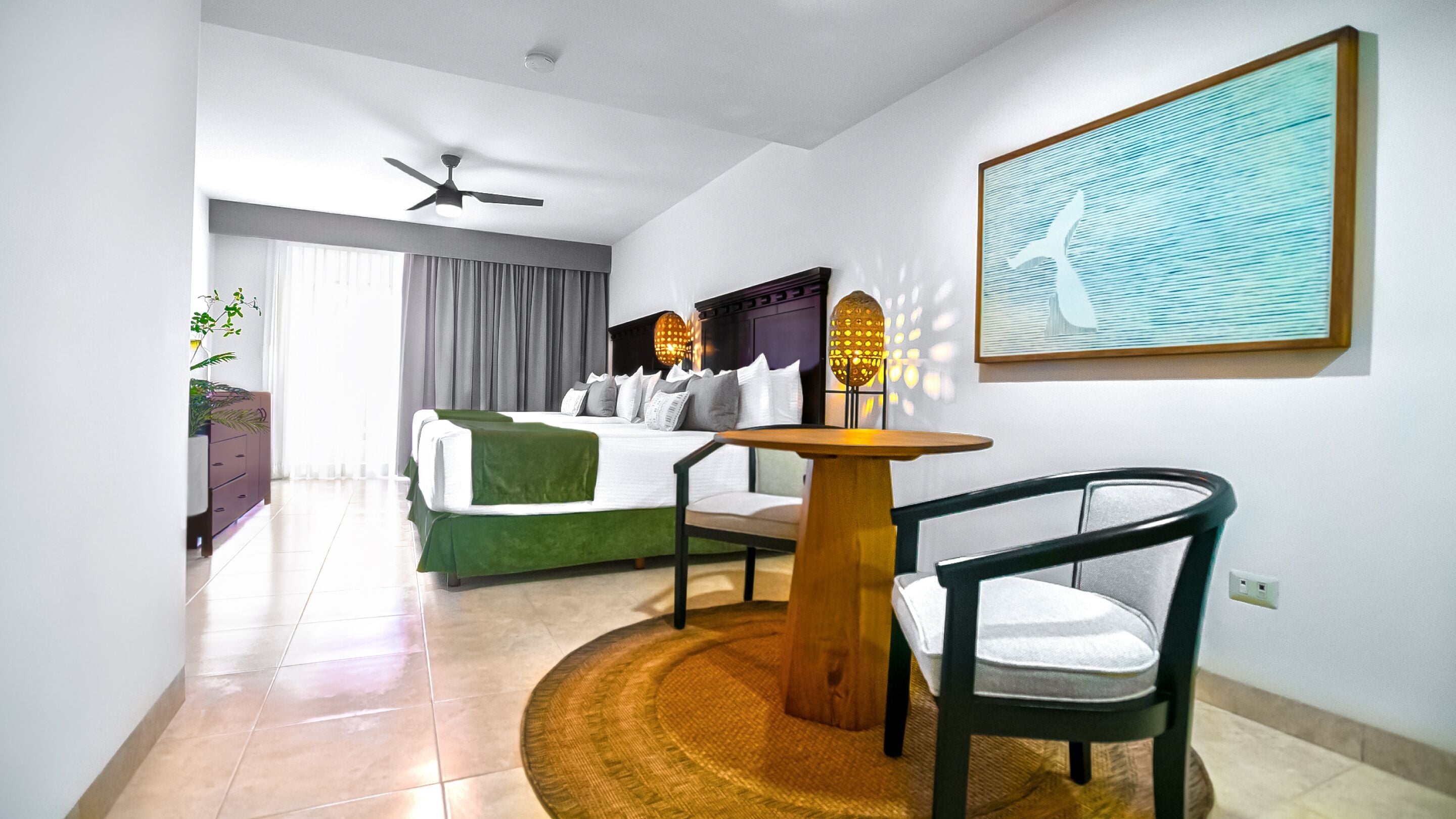 A hotel room with a bed, a table, a chair, a ceiling fan, and a wall-mounted light fixture.