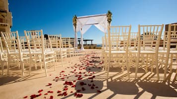 Un lieu de réception en plein air pour un mariage, avec des chaises blanches disposées en rangées, une arche blanche décorée de fleurs et un ciel bleu azur.
