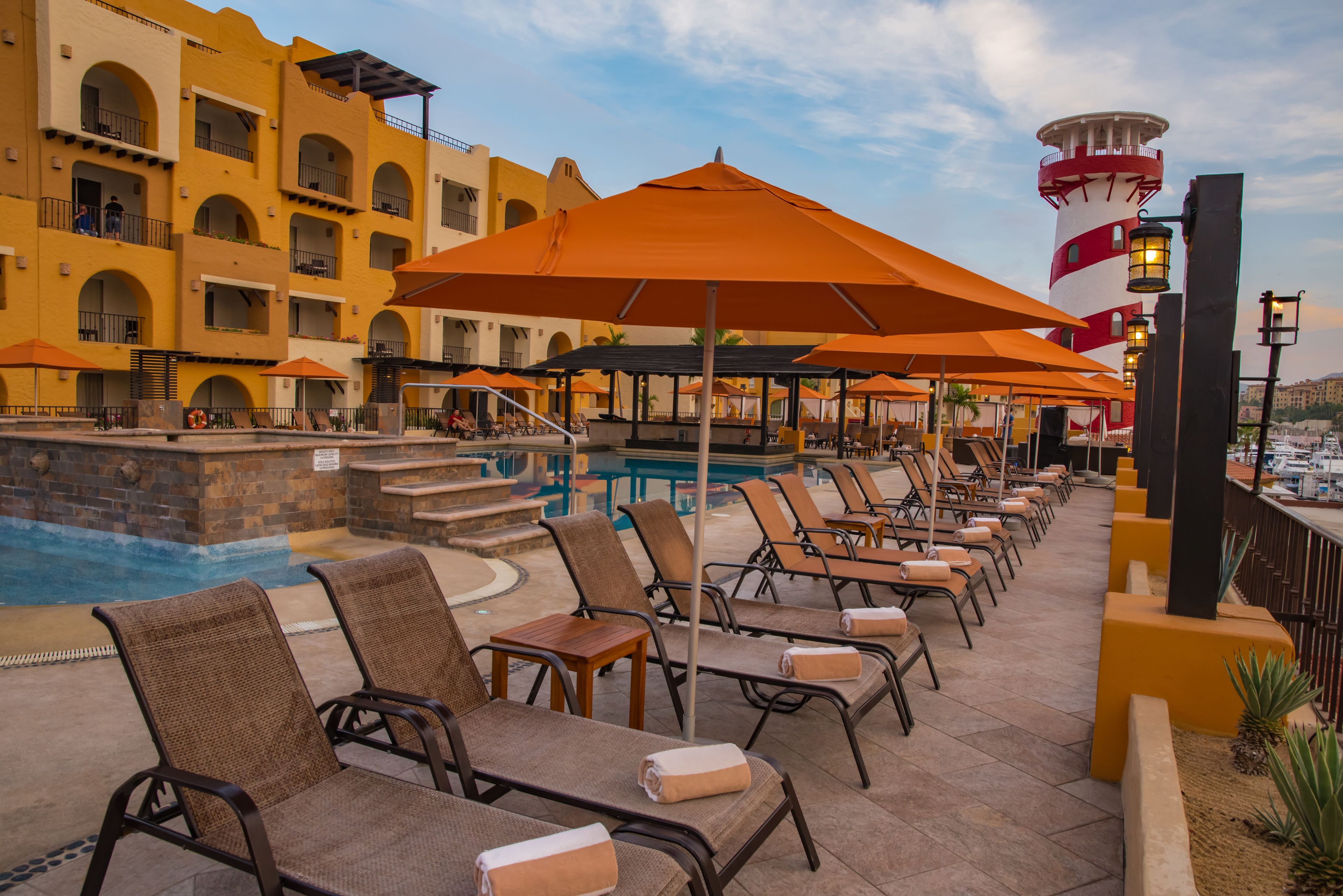A poolside area with lounge chairs and umbrellas, a pool, and a building with arched balconies.