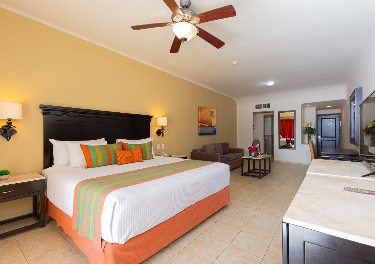 Habitación de hotel con una cama grande, ventilador de techo, escritorio, silla, televisor y kitchenette.