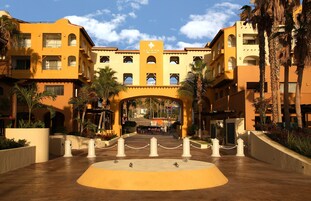 Una entrada de hotel con un arco central, palmeras y cielo despejado.