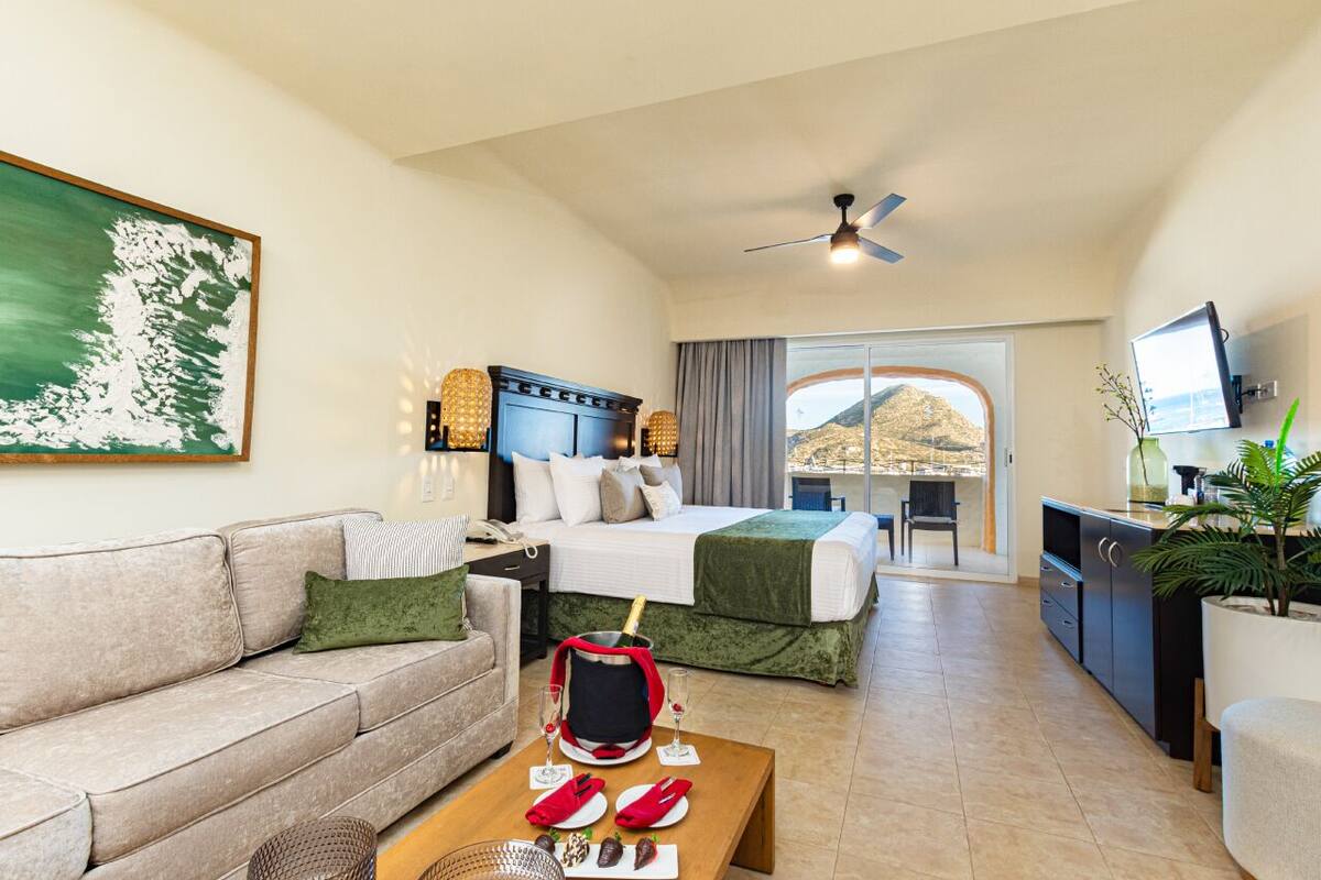 Habitación de hotel con cama, sofá, mesa de comedor y una botella de champán, y televisor.