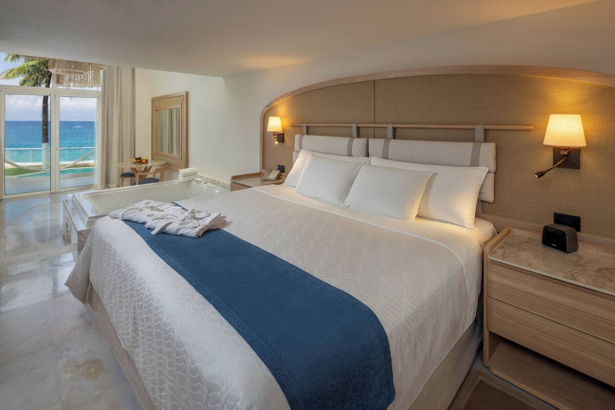 A hotel room with a large bed, a desk, a chair, a nightstand, and a view of the ocean.