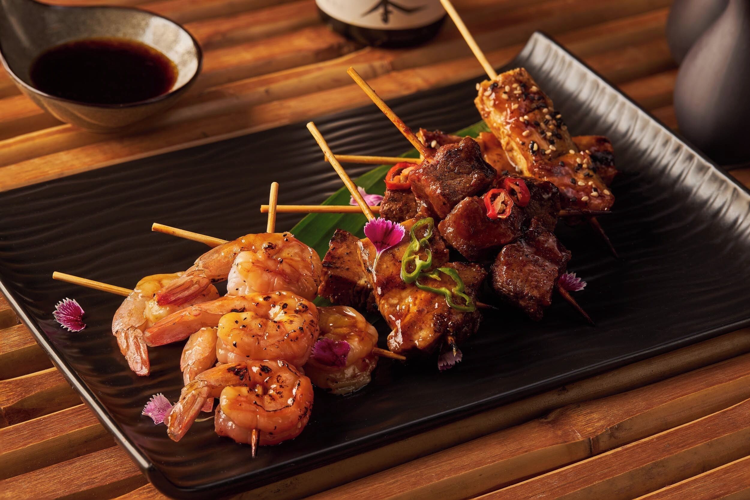 A plate of skewered shrimp and meat, garnished with sesame seeds and chili, served on a black rectangular plate.