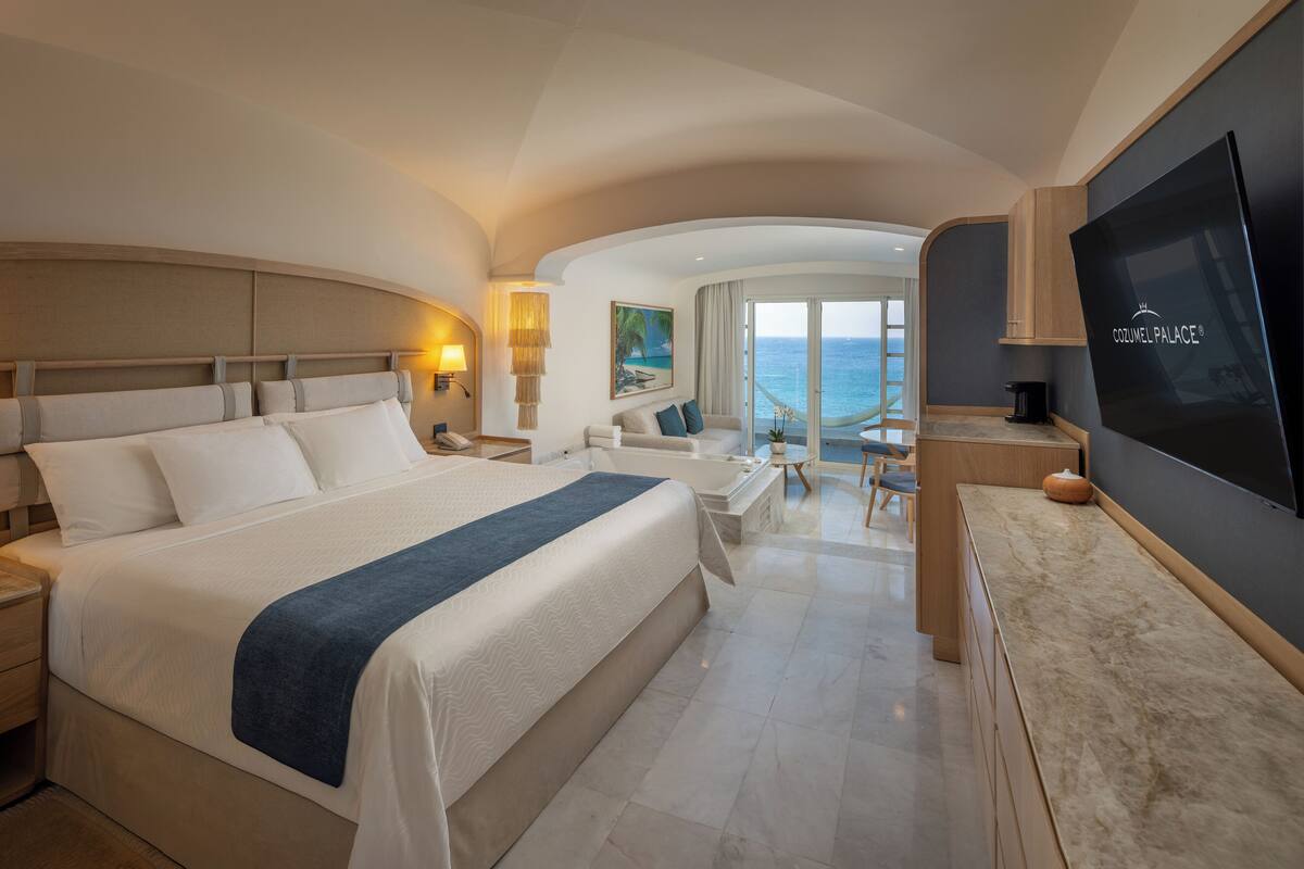 A modern hotel room with a large bed, a TV, a balcony with a view of the sea, and a dining area.